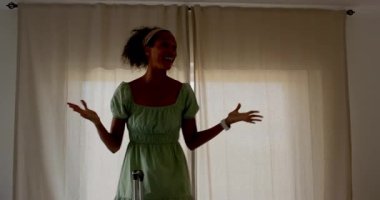 Yeşil elbiseli kadın pencerenin önünde, yanında bavuluyla neşeyle dans ediyor. Seyahat, tatil, özgürlük, mutluluk, kutlama, hareket