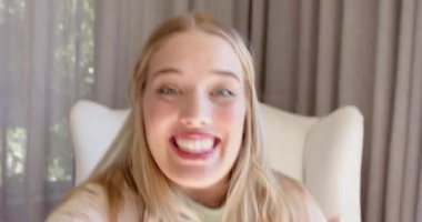 Kadın içerik yaratıcısı izleyicileri selamlıyor, evdeki arka koltukta oturuyor, perdelerin yanında el kol hareketi yapıyor. Portre, yaşam tarzı, hoş geldin, rahat, doğal ışık, iletişim, moda