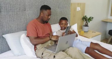 Afro-Amerikalı baba ve oğul laptopu yatağın üzerine koyuyorlar tablete vuruyorlar ve baba işaret ediyor ve gülüyor. Aile, bağ, teknoloji, rahatlama, ev, dijital, bağlantı