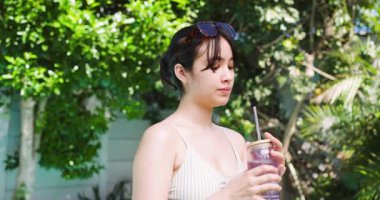 Mayo giymiş Asyalı bir kadın, güneşli bahçede gözleri gölgeliyor, bardaktan içiyor, güneş gözlüğünü ayarlıyor. Doğa, rahatlık, ferahlık, yaşam tarzı, canlılık, sağlık, açık hava.