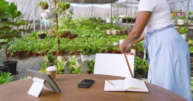 Afro-Amerikalı kadın serada defterine yazıyor. Etrafı bitkilerle çevrili. Bahçıvanlık, bahçıvanlık, yeşillik, not alma, bitki örtüsü, etnik