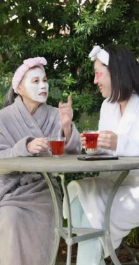 Dikey video: Kadınlar dışarıda çay içerken yüz maskeleri ve bornozlar takıyor, birlikte dinleniyorlar. Rahatlama, sağIık, arkadaşlık, bağlanma, sükunet, güvenlik.