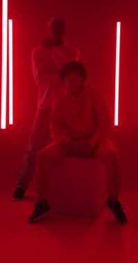 Dikey video: Kapüşonlu genç adam kırmızı neon ışıkların altında oturmuş, dalgın dalgın bakıyor. Sokak giyimi, kentsel, fütürist, tefekkür, moda, modern