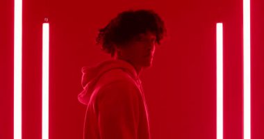 Kırmızı ışıklar yanarken, erkek kapüşonlu bir kapüşonlu giyip neon kaplamalı stüdyoda dans pozu veriyor. Stüdyo, neon, dans, sanatçı, minimalist, ruh hali, çağdaş