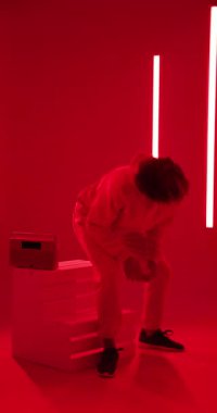 Dikey video: Kapüşonlu genç adam kırmızı ışıklı stüdyoda kutuda oturuyor, dalgın dalgın bakıyor. Düşünce, moda, karamsar, kentsel, iç gözlem, modern