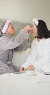 Dikey video: Anne ve kızı birlikte cilt maskesi takıyor, rahat bornozlar giyiyorlar. Rahatlama, sağlık, aile, gençleşme, kendine bakım, şımartma.