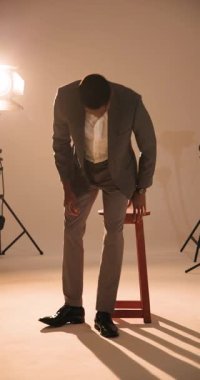 Dikey video: Kendine güvenen şık takım elbiseli adam profesyonel fotoğraf stüdyosundaki taburede poz veriyor. Moda, portre, zarafet, özgüven, erkek giyim, model