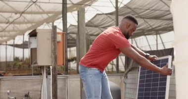 Inspecting solar panel in greenhouse, African man focusing on sustainable energy solutions. Renewable, technology, eco-friendly, inspection, innovation, power