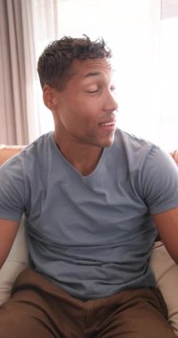 Dikey video: Sıradan giyinmiş gülümseyen adam evdeki koltukta oturmuş kameraya bakıyor. Yaşam tarzı, rahatlama, evcil, boş zaman, portre, konfor