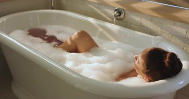 Güneş ışığı jaluzilerden süzülüyor. Kadın küvette sırılsıklam oluyor. Dizlerini büküyor ve baloncuk çıkartıyor. Lüks, rahatlama, minimalist, sağIık, sükunet, kişisel bakım, spa