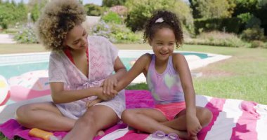 African American mom and daughter applying orange sunscreen on towel beside pool for pool play. Family, outdoor, leisure, summer, wellness, bonding, recreational