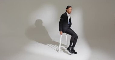 Asyalı erkek manken kayıyor, ayakta duruyor, ceketini stüdyo ışıkları altında çekimler için tabureye asıyor. Moda, zarafet, profesyonellik, portre, minimalizm, erkek, stüdyo