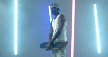 Afrikalı Amerikalı adam neon ışıklı stüdyoya giriyor, şapkasını ayarlıyor, kaykay sallıyor, gösteriş yapıyor. Kentsel, modern, enerjik, enerjik, yaşam tarzı, moda