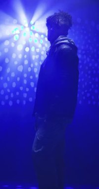 Dikey video: Kapüşonlu genç adam kasvetli stüdyo atmosferinde mavi ışıkların altında duruyor. Moda, kentsel, dramatik, aydınlık, portre, gündelik