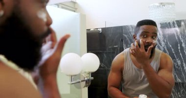 Afrika kökenli Amerikalı bir adam modern banyoda dikkatli bakım için ayna kullanarak yüz kremi sürüyor. Tımarlama, yansıtma, cilt bakımı, modern, banyo, özgüven.