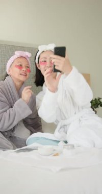 Dikey video: Anne ve kızı yatak odasında akıllı telefona bakarken bornozla gülüyorlar. Aile, bağ, teknoloji, eğlence, lüks, rahatlama