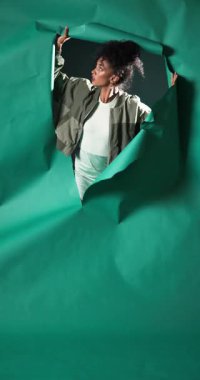 Vertical video: Woman tearing through green paper backdrop, expressing curiosity and exploration. adventure, innovation, breakthrough, creativity, discovery, mystery