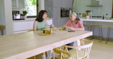 Asyalı anne ve kızı bardaklarını tokuşturup çay dolduruyor salata karışımını mutfak masasında paylaşıyorlar. Aile, bağ, mutfak, aşçılık, modern, neşeli, iletişim