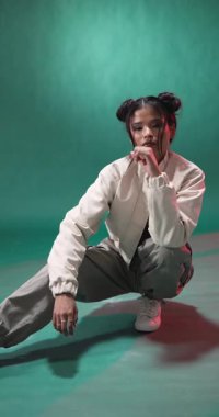 Dikey video: Gündelik kıyafetli genç bir kadın şık stüdyo ortamında güven içinde çömeliyor. Moda, çağdaş, trend, şehir, poz, tutum