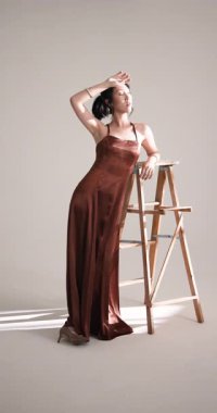 Dikey video: Saten elbiseli zarif bir kadın stüdyoda ahşap merdivene karşı düşünceli bir şekilde poz veriyor. Moda, zarafet, sofistike, tarz, cazibe, lüks