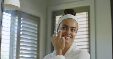 Krem sürdükten sonra banyo aynasında bornoz giyen kadın yanaklarına masaj yapıyor ve gülümsüyor. Cilt bakımı, rahatlama, kendine bakım, güzellik, ev, sağlık, minimalizm.