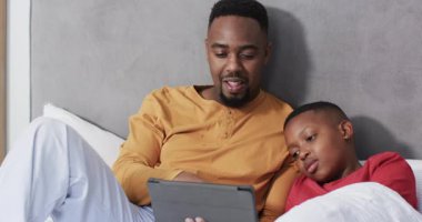 Afro-Amerikalı baba ve oğul tablet tıkırdatıyor, oğlu yatakta şaşkınlığını paylaşıyor. Aile, bağ, rahatlama, samimiyet, teknoloji, konforlu, hassas