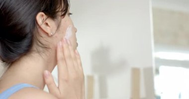 Asyalı kadın krema kavanozunu kıvırıyor parmağını daldırıyor ve banyo süslemesinde yüzünü nemlendirmek için masaj yapıyor. Cilt bakımı, güzellik, bencillik, susuzluk, refah, minimalizm, doğal ışık