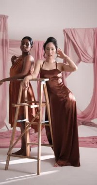 Dikey video: Moda stüdyosunda merdivenle poz veren zarif kahverengi elbiseli modeller. zerafet, cazibe, tarz, giysi, moda, moda