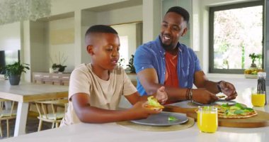 Pizza geldikten sonra Afrika kökenli Amerikalı baba ve oğul mutfak adasında yemek yiyor, beşlik çakıyor. Aileler, kaynaşma, mutfak, çağdaş, sıcak, mutluluk, etkileşim