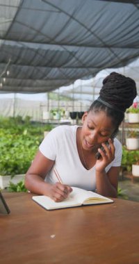 Dikey video: Serada gülümseyen kadın telefonla konuşuyor ve deftere yazıyor. Bahçıvanlık, iletişim, çoklu görev, bahçıvanlık, iş, teknoloji