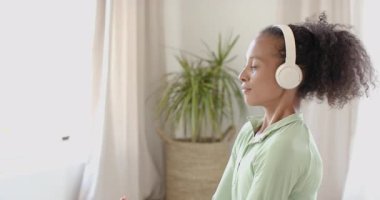 Genç bir kadın kulaklıkla meditasyon yapıyor, evdeki farkındalığa odaklanıyor. Meditasyon, rahatlama, zen, sükunet, barış, yaşam tarzı