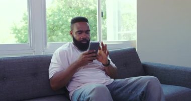 Parlak odada gri koltukta oturan Afro-Amerikalı adam, açık pencere manzaralı akıllı telefonu kontrol ediyor. Hüsran, tefekkür, akıllı telefon, rahat, modern, doğal ışık, sükunet.