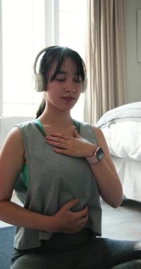 Dikey video: Asyalı kadın kulaklıkla meditasyon yapıyor, evde nefes egzersizlerine odaklanıyor. Meditasyon, rahatlama, farkındalık, sağIık, sükunet, kişisel bakım.