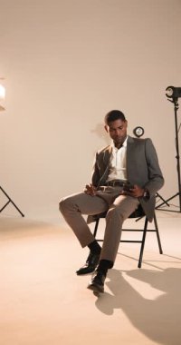 Vertical video: Thoughtful man in stylish suit sitting on chair in professional studio. Fashion, elegance, sophistication, portrait, luxury, modern
