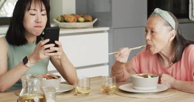 Asyalı Çinli anne ve kız ısırık arası veriyor. Mutfakta telefon özçekimini paylaşıyor ve çay kızartıyor. Aile, nesiller arası bağ, teknoloji, yemek, yaşam tarzı, samimiyet