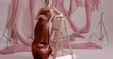 Asyalı model stüdyodaki kameraya yanıt veriyor, tahta merdivene tırmanıyor ve saten elbiseler giyiyor. Zarafet, duruş, moda, cazibe, minimalist, sofistike, portre