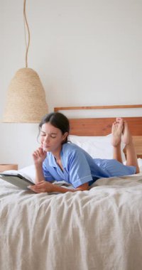 Dikey video: Evde dinlenme, pijamalı bir kadın yatakta kitap okuyor, boş zamanların tadını çıkarıyor. Rahatlama, konfor, uyku zamanı, rahatlık.