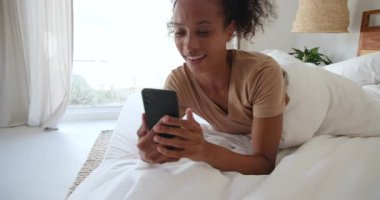 Akıllı telefon kullanarak yatakta gülümseyen kadın, evde sabah rahatlığının tadını çıkarıyor. Teknoloji, yaşam tarzı, rahatlık, rahatlık