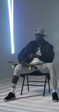 Dikey video: Sandalyede oturan genç adam kaykay tutuyor, sokak kıyafetleri giyiyor. Kaykaycı, moda, şehir, yaşam tarzı, milenyum, giyim