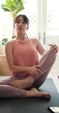 Dikey video: Asyalı kadın evde yoga minderinde smoothie ve akıllı telefonuyla dinleniyor. Sağlık, meditasyon, rahatlama, dijital, yaşam tarzı, besleyici