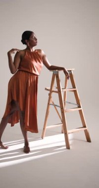 Dikey video: Şık elbiseli zarif kadın stüdyoda tahta merdivenle güven içinde poz veriyor. Moda, zarafet, tarz, güven, portre, modellik