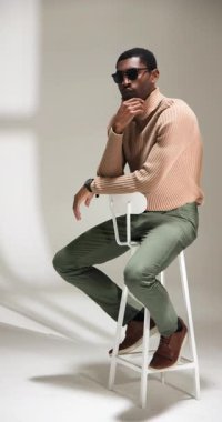 Dikey video: Güneş gözlüklü şık adam modern stüdyodaki taburede güven içinde poz veriyor. Moda, güven, portre, gözlük, tarz, moda