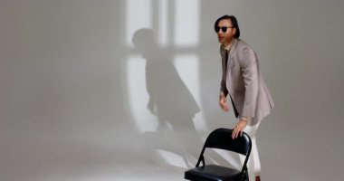 Asyalı erkek, elinde güneş gözlüğü ile stüdyoya giriyor. Pencere ışığını kontrol ediyor. Sandalyeye oturuyor. Şık, çağdaş, minimalist, profesyonel, çok yönlü, modaya uygun, dinamik