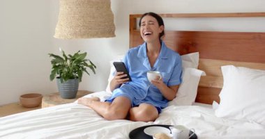 Yatakta dinleniyor, pijamalı bir kadın kahve içiyor ve telefonda konuşuyor. Rahatlama, konfor, yaşam tarzı, rahatlık, sohbet, sabah