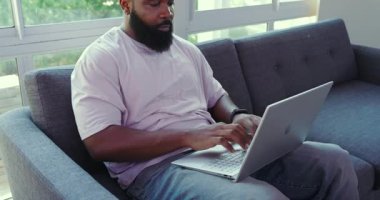 Kanepede oturan Afro-Amerikalı adam yeşil manzaralı kapalı alanda dizüstü bilgisayarla çalışıyor. Laptop, gündelik, yeşillik, çalışma alanı, üretkenlik, sükunet, rahatlama