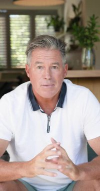 Dikey video: Normal giyinen yetişkin bir adam evde oturuyor, düşünceli ve rahat görünüyor. Yaşam tarzı, tefekkür, yetişkin, boş zaman, huzur, ev