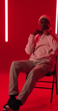 Dikey video: Kapüşonlu genç adam canlı stüdyo ışıklarıyla koltukta güvenle oturuyor. Moda, güven, modern, kentsel, tarz, erkek