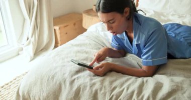 Evde dinleniyor, pijamalı bir kadın rahat bir yatakta akıllı telefon kullanıyor. Rahatlama, rahatlama, teknoloji, konfor, yaşam tarzı, dijital