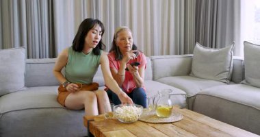 Uzaktan kumandaya basan yaşlı Çinli kadın, patlamış mısır ve yastık sunan kız, kanepede abur cubur yiyen. Aile, bağ, rahatlama, ev, rahatlık, sıcaklık, birliktelik.