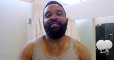 Beyaz bornozla parlak banyoya sakal yağı süren Afro-Amerikalı bir adam. Bakım, rahatlama, sağIık, güven, tazelik, minimalizm, ışıklandırma